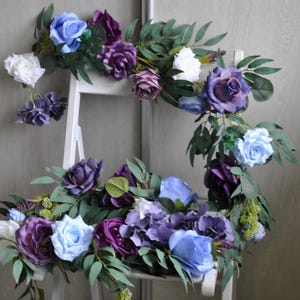 Può includere: Una composizione floreale con rose e ortensie artificiali nelle tonalità azzurro, viola intenso e bianco. I fiori sono intervallati da foglie di eucalipto verdi e fogliame, creando un'esposizione botanica a cascata.