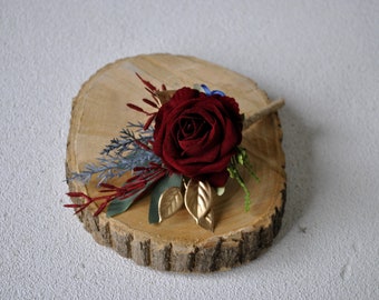 May include: A burgundy rose boutonniere with gold leaves, blue and red sprigs, and a wooden stem. The boutonniere is displayed on a wooden slice.