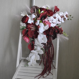 May include: A cascading floral arrangement featuring a mix of white orchids, burgundy calla lilies, and deep red roses. The bouquet includes accents of pink and white flowers, with trailing burgundy beaded strands. The arrangement is displayed on a white slatted chair.