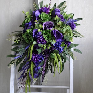 May include: A cascading bouquet of artificial flowers in shades of purple and green. The arrangement includes roses, hydrangeas, and foliage. The bouquet is set against a white backdrop.