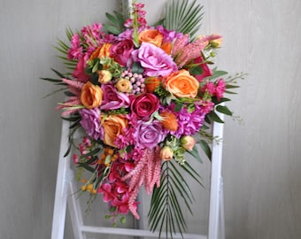 May include: A vibrant floral arrangement featuring a cascade of colorful flowers. The bouquet includes orange, pink, and purple roses, along with other blooms and greenery. The flowers are arranged on a white structure, creating a visually appealing display.