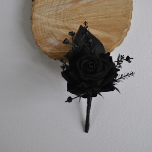 May include: A black rose boutonniere with black leaves and berries. The boutonniere is on a wooden background.