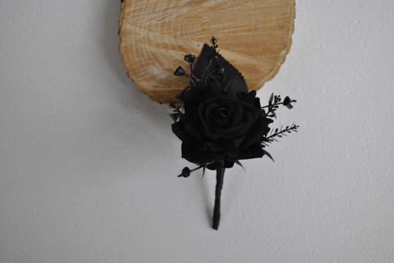 May include: A black rose boutonniere with black leaves and berries. The boutonniere is on a wooden background.
