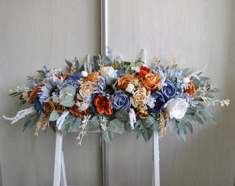 May include: A horizontal floral arrangement featuring a mix of artificial roses and peonies in shades of burnt orange, dusty blue, navy, and cream. The bouquet includes various green eucalyptus leaves, white filler flowers, and wispy dried-style stems.