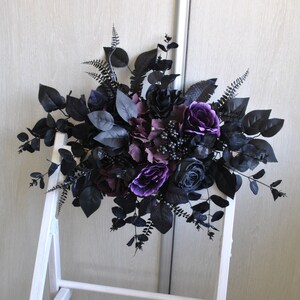 May include: A black and purple floral arrangement with roses, berries, and greenery. The arrangement is displayed on a white easel.