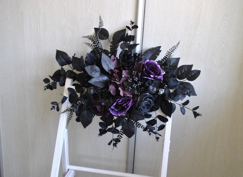 May include: A black and purple floral arrangement with roses, berries, and greenery. The arrangement is displayed on a white easel.
