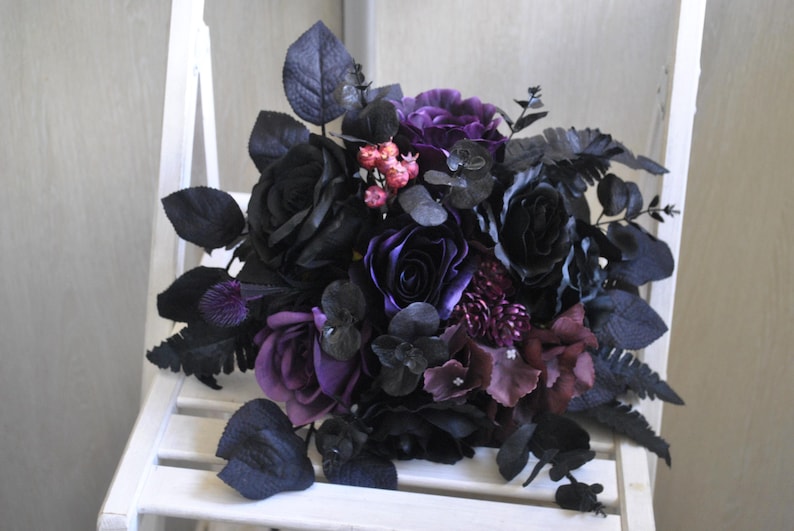 May include: A dark purple and black bridal bouquet with roses, ferns, and other foliage.