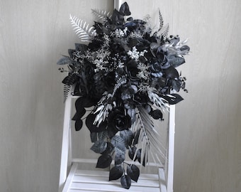 May include: A cascading floral arrangement featuring black roses, leaves, and silver accents. The bouquet is displayed on a white chair, creating a striking contrast. The arrangement includes various textures and shapes, suitable for home decor or special events.