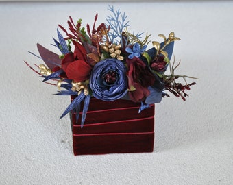 May include: A small, red velvet box with a floral arrangement. The flowers are blue, red, and gold. The arrangement is made up of various types of flowers, including roses, carnations, and sprigs of greenery.