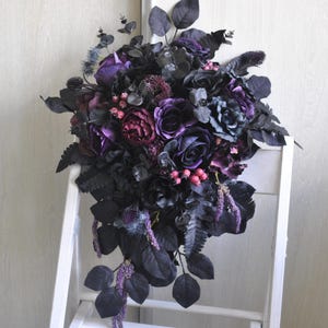 May include: A cascading bouquet of dark purple and black silk flowers with green leaves. The bouquet is arranged in a teardrop shape and is perfect for a gothic wedding or event.