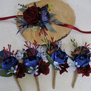 May include: A collection of floral wedding accessories featuring deep red and blue flowers. The set includes a wrist corsage with a red ribbon and several boutonnieres with twine-wrapped stems, arranged on a wooden slice.
