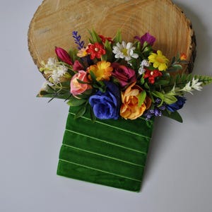 Op de afbeelding: Een groene fluwelen doos met een boeket gekleurde kunstbloemen, waaronder rode, gele, blauwe en roze bloemen.