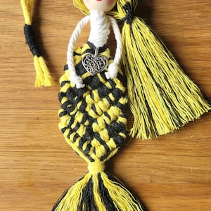 Könnte beinhalten: Eine gelbe und schwarze Garn-Meerjungfrauenpuppe mit einem weißen Garn-Oberteil und einem silbernen Herz-Anhänger. Die Puppe hat einen Holzkopf mit bemalten Augen und einem Lächeln.