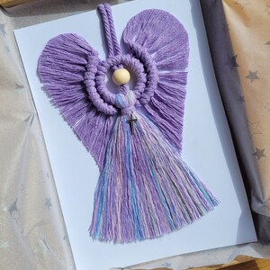 May include: A purple macrame angel with a wooden bead and a silver cross hanging from the bottom. The angel has a heart-shaped body and fringed wings.