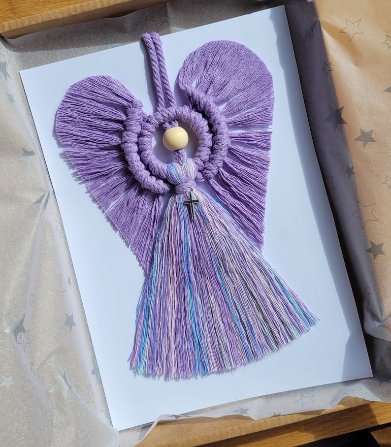 May include: A purple macrame angel with a wooden bead and a silver cross hanging from the bottom. The angel has a heart-shaped body and fringed wings.