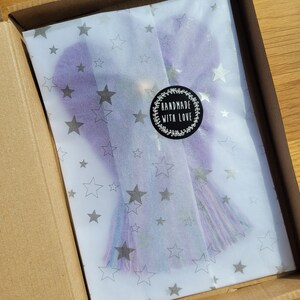 May include: A purple and blue tassel with silver glitter is wrapped in white tissue paper with silver stars. The tissue paper is inside a brown cardboard box. A black and white sticker with the text "Handmade with Love" is on the tissue paper.