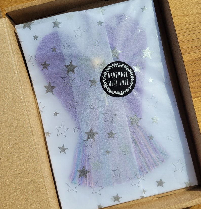 May include: A purple and blue tassel with silver glitter is wrapped in white tissue paper with silver stars. The tissue paper is inside a brown cardboard box. A black and white sticker with the text "Handmade with Love" is on the tissue paper.
