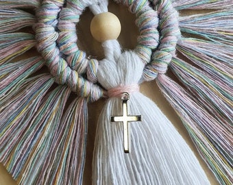 May include: Macrame angel wall hanging with a silver cross pendant. The angel features a circular halo with pastel-colored accents, a wooden bead, and a white tassel. The wings are made of pastel-colored strands.