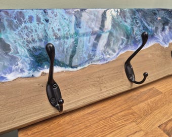 May include: A wooden wall-mounted coat rack featuring a natural light brown wood base and a glossy resin top layer with swirling blue, teal, and white patterns. Three black metal hooks are attached to the front of the wooden board.