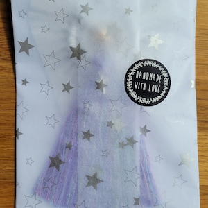 May include: A purple and blue tassel with a silver star pattern on a white background. The tassel is packaged in a white tissue paper envelope with a black circle sticker that says "Handmade with Love".