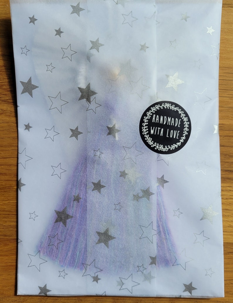 May include: A purple and blue tassel with a silver star pattern on a white background. The tassel is packaged in a white tissue paper envelope with a black circle sticker that says "Handmade with Love".