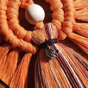 May include: Orange and white macrame wall hanging with a wooden bead and a pumpkin charm. The macrame is made with a variety of knots and has a fringed bottom.