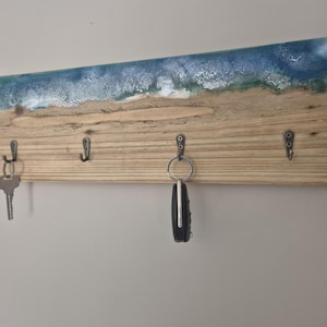 May include: Wooden key holder with a beach-themed design. The top has a blue and white resin wave pattern, transitioning to a sandy beach colour. Four metal hooks are attached for keys. Keys and a car key fob hang from the hooks.
