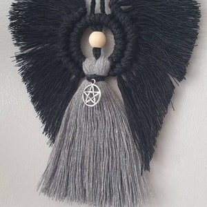 May include: A gray and black macrame angel with a pentagram charm hanging from its waist. The angel has a wooden bead head and black fringe wings.