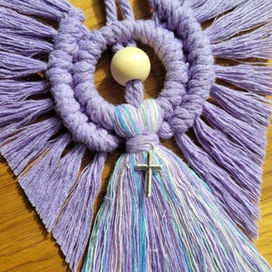 May include: A purple and white macrame wall hanging with a wooden bead and a silver cross charm. The macrame is made with a variety of knots and textures.