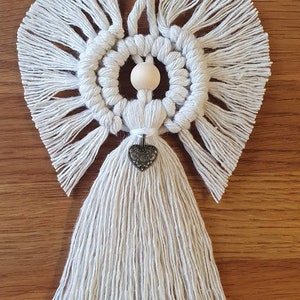May include: A white macrame angel ornament with a wooden bead head and a silver heart charm. The angel has long, flowing wings and a tassel skirt.