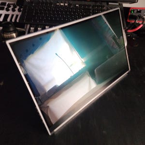 Könnte beinhalten: Ein rechteckiger Computermonitor mit weißem Rahmen. Der Bildschirm spiegelt die Umgebung wider, einschließlich einer Tastatur und anderer elektronischer Geräte. Der Monitor befindet sich auf einer dunklen Oberfläche.