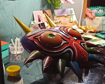 Puede incluir: Una réplica pintada a mano de la Máscara de Majora, con detalles intrincados. La máscara está hecha de un material de color morado oscuro, rojo y verde con un contorno negro. La máscara tiene un ojo grande, rojo con un iris verde y un ojo negro más pequeño con un iris blanco. La máscara está sentada sobre una superficie negra.