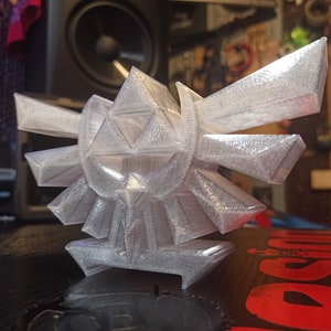 May include: A white 3D printed model of the Triforce symbol from the Legend of Zelda video game series. The model is detailed and intricate, with sharp edges and a glossy finish.