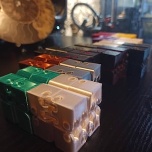 May include: A set of colorful 3D printed fidget toys, stacked in a row. The toys are made of different colors, including green, white, silver, copper, and black. The toys are all the same size and shape, and they have a unique design that allows them to be flipped and rotated.