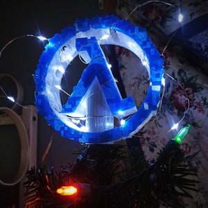 May include: A blue and white Christmas tree topper featuring the Half-Life logo. The logo is surrounded by white lights.