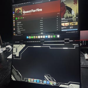 May include: A black laptop with a second screen above it, displaying a music streaming service with the album "Quest For Fire" by Skrillex. The laptop's screen shows a black and grey design with app icons.