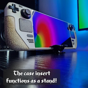 May include: A white and gray handheld gaming device with a rainbow-colored screen. The device has a textured grip and a black stand. The text "The case insert functions as a stand!" is displayed at the bottom.
