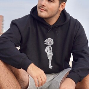 May include: A person wearing a black hooded sweatshirt with a white graphic of a person with a fish head. The person is sitting on a rock.