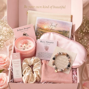 May include: A gift box containing a pink candle with crystals, a beaded bracelet with a gold charm, a satin scrunchie, a pink sleep mask, a small pouch, and informational cards labelled Zebra Jasper and Rose Quartz, with the text Be your own kind of beautiful.