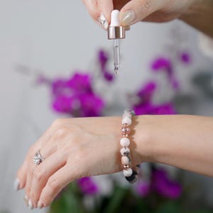 May include: A beaded bracelet featuring round, mottled pink and white stones interspersed with metallic rose gold-toned spherical spacers. The bracelet is worn on a wrist while a clear glass dropper with a rose gold collar is held above it.