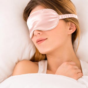 May include: A pale pink sleep mask with a smooth, satin-like finish and a matching gathered elastic strap. The mask features a contoured shape designed to cover the eyes, with visible piping along the edges and a soft, uniform light pink colour.