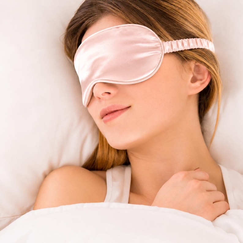 May include: A pale pink sleep mask with a smooth, satin-like finish and a matching gathered elastic strap. The mask features a contoured shape designed to cover the eyes, with visible piping along the edges and a soft, uniform light pink colour.