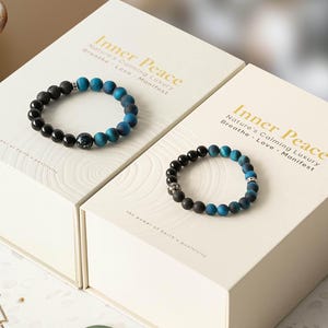 May include: Two bracelets with black and blue beads are displayed on white boxes. The boxes have the text "Inner Peace" and "Nature's Calming Luxury, Breathe, Love, Manifest". The bracelets are made of various sizes of round beads.