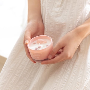 May include: A light pink candle in a matching pink glass container with white wax. The candle features a single centred wick and is topped with several small, light pink crystal-like stones embedded in the surface of the wax.
