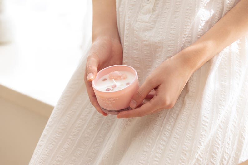 May include: A light pink candle in a matching pink glass container with white wax. The candle features a single centred wick and is topped with several small, light pink crystal-like stones embedded in the surface of the wax.