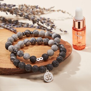 May include: Two beaded bracelets with grey and black stones, one with a silver infinity charm and the other with an Om symbol charm. A small bottle of oil with the text "Seilla Inner Peace" is also present.