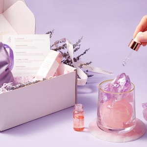 May include: A pink and white gift box containing a lavender scent card and a small bottle of essential oil. The box is filled with lavender-coloured tissue paper. A glass jar with a gold rim holds amethyst crystals and a pink candle. The candle has a gold label with the word "Soulstone" on it.