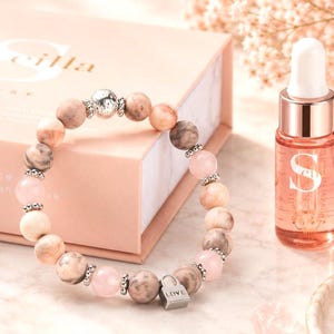 May include: A beaded bracelet featuring alternating round pink and marbled beige stones, accented with small silver-toned spacers and a central silver-toned padlock charm engraved with the word LOVE, positioned near a small glass bottle of oil.
