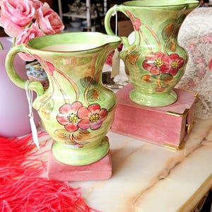 May include: Two green ceramic jugs with floral designs. The jugs have a light green glaze with pink and yellow flowers. The jugs are sitting on pink velvet boxes.