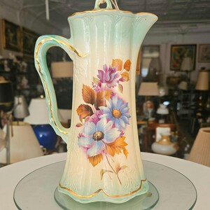 Peut inclure: Un pichet en porcelaine vintage avec un motif floral bleu clair et blanc. Le pichet a une bordure dorée et une poignée. Il est posé sur une assiette en verre transparent.